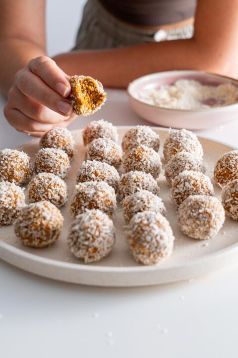 Hand holding a half-bitten Carrot Cake Bliss Ball with the plate in the background.