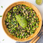 Overhead view of edamame and forbidden rice salad in a wooden bowl with sliced avocado on top, served with wooden salad servers and a halved lime on a white surface