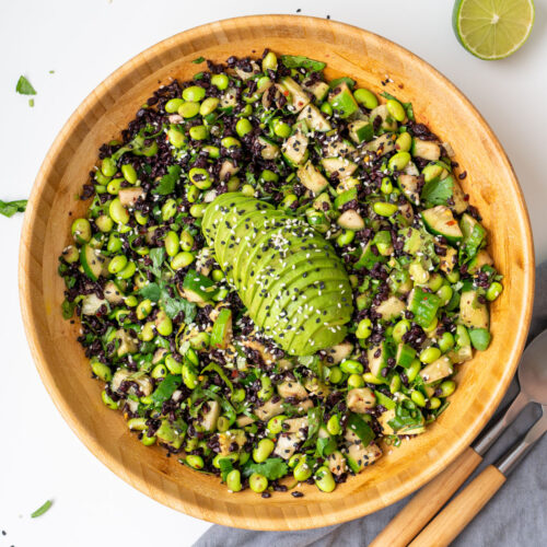 Overhead view of edamame and forbidden rice salad in a wooden bowl with sliced avocado on top, served with wooden salad servers and a halved lime on a white surface