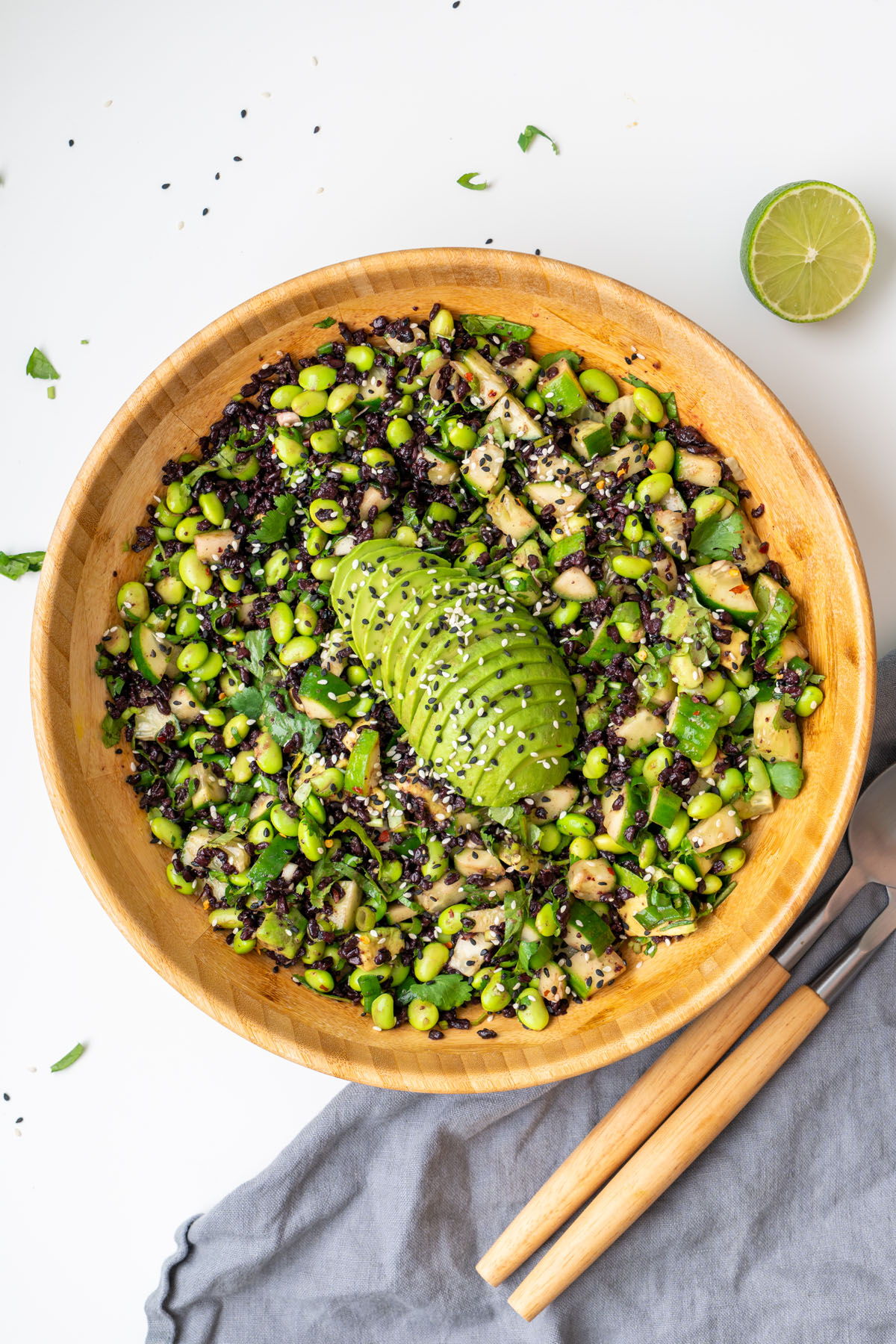 Overhead view of edamame and forbidden rice salad in a wooden bowl with sliced avocado on top, served with wooden salad servers and a halved lime on a white surface