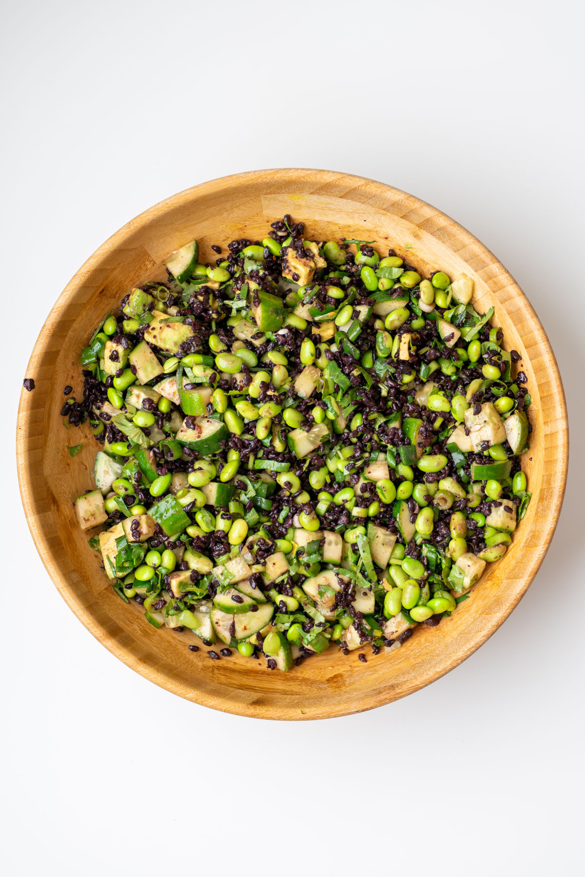 Overhead view of edamame and forbidden rice salad fully tossed in a wooden bowl, showing black rice, edamame, cucumber, and avocado coated in dressing