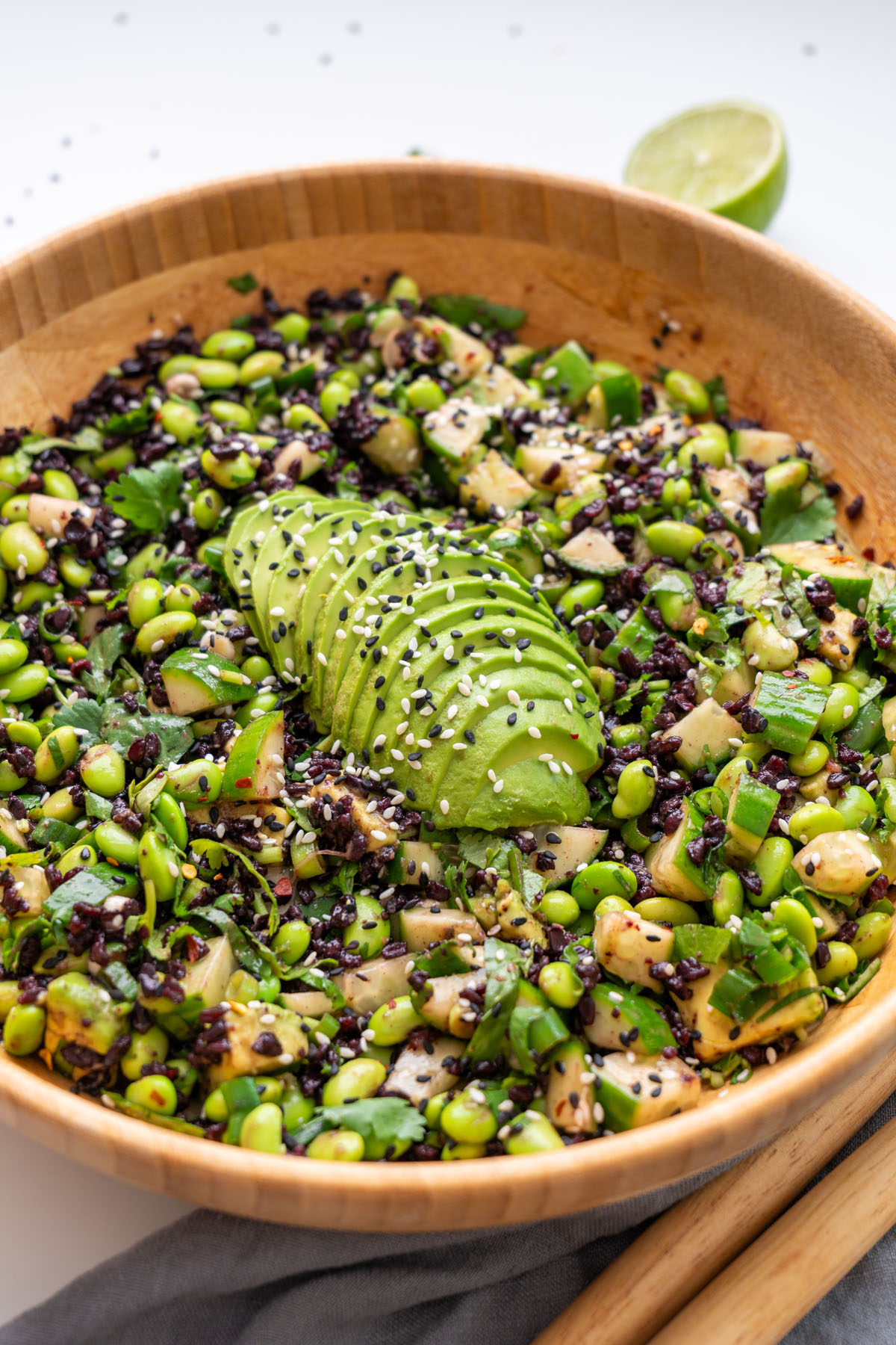 Angled close-up of edamame and forbidden rice salad in a wooden bowl, with fanned sliced avocado topped with black and white sesame seeds and a lime in the background