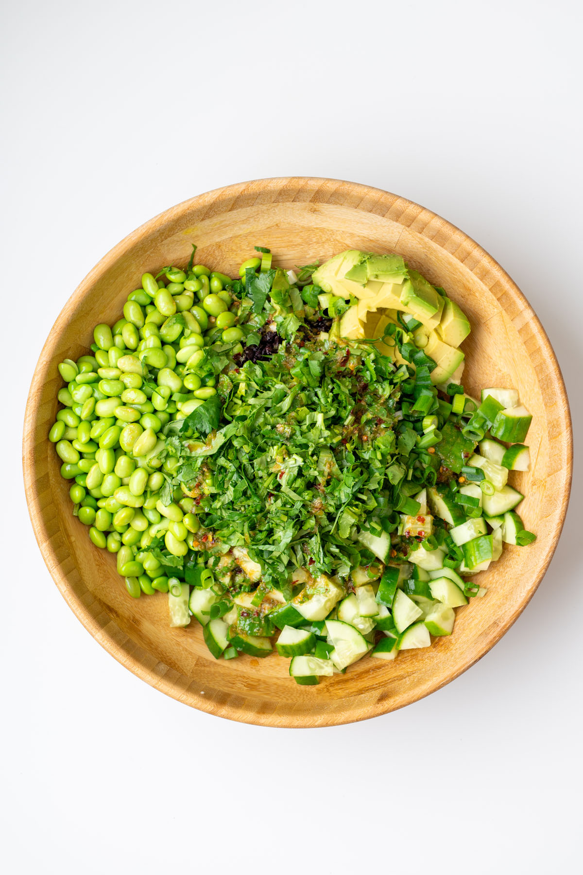 Overhead view of fresh salad ingredients in a wooden bowl before mixing, including edamame, diced cucumber, diced avocado, chopped cilantro, and dressing poured over