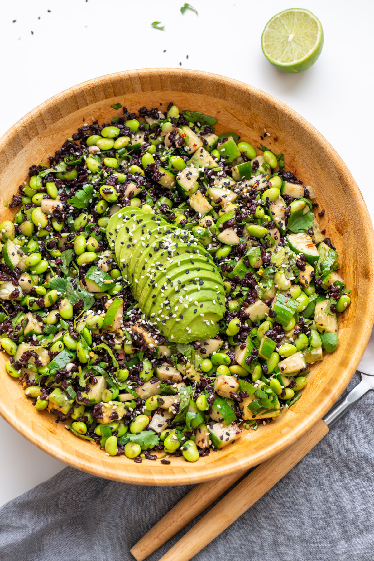 Overhead shot of edamame and forbidden rice salad in a wooden bowl topped with sliced avocado and sesame seeds, with salad servers and a halved lime on a white surface