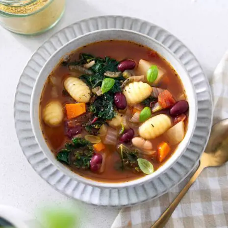 A bowl of hearty minestrone soup filled with vegetables, beans, and pasta, topped with a sprinkle of vegan cheese.