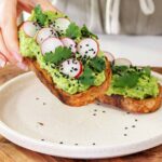 Sarah holding a Smashed peas and avocado toast on a white plate on a wooden serving board