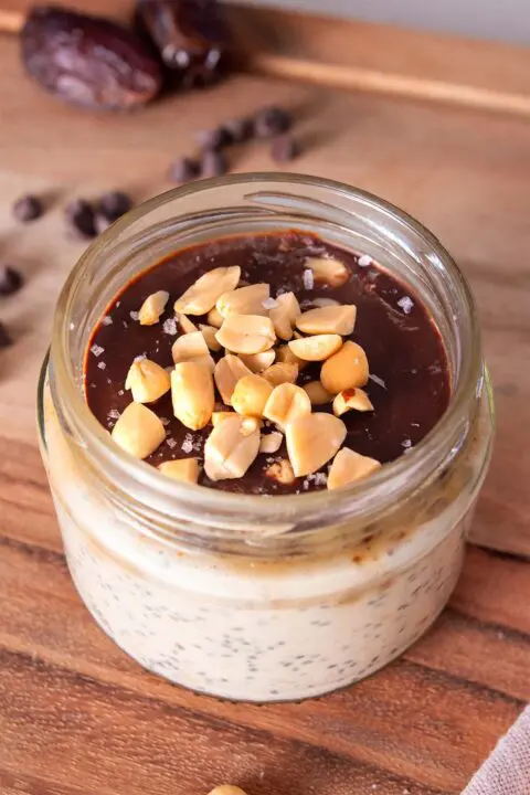 Jar with layered overnight oats, including oats, peanut butter, and chocolate, topped with peanuts and sea salt, on wood.
