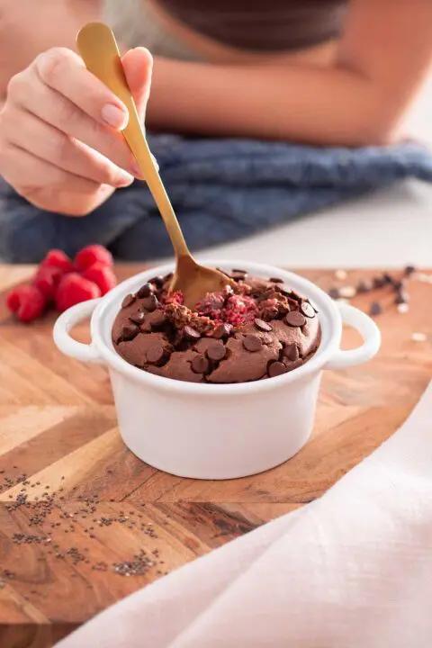 A close-up of chocolate raspberry baked oats in a white ceramic dish, topped with chocolate chips and a spoonful being taken, revealing the moist interior.