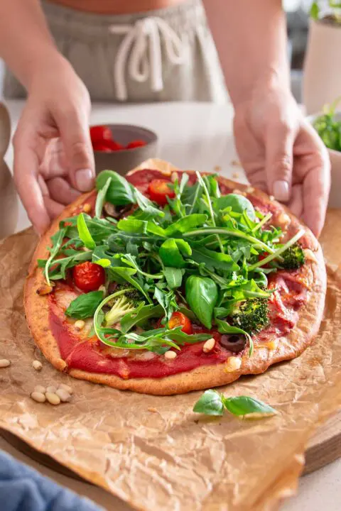 Hands delicately placing fresh arugula and herbs on a baked gluten-free pizza crust, showcasing the process of adding fresh, leafy greens to enhance the flavors of the vegan pizza.