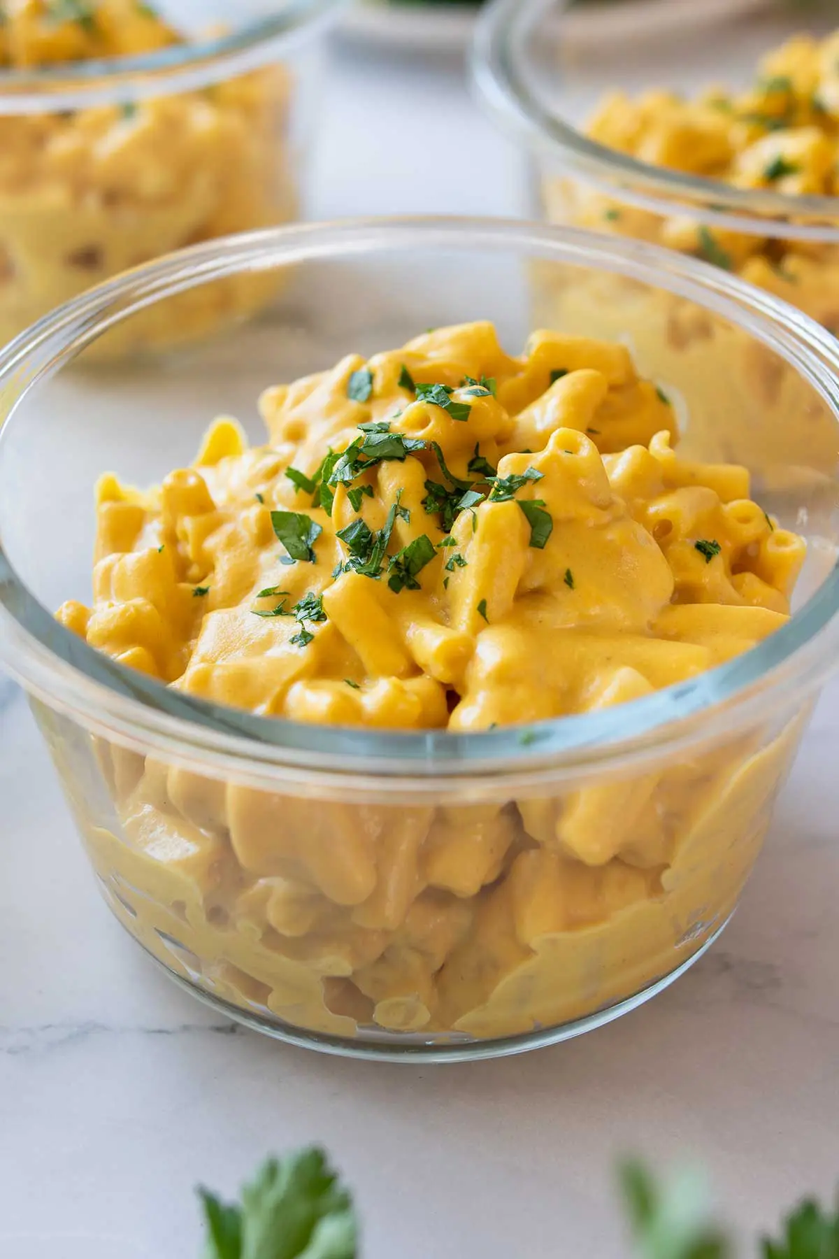 Close-up view of a glass bowl with vegan high-protein mac and cheese garnished with parsley on a marble surface.