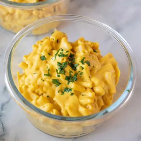 Two glass bowls on a marble counter filled with creamy vegan high-protein mac and cheese, topped with chopped fresh parsley.