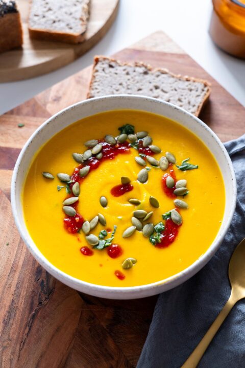 Closeup of a healing bowl of smooth butternut squash and red lentil soup garnished with seeds and red sauce, accompanied by slices of bread and a gold spoon on a rustic board.