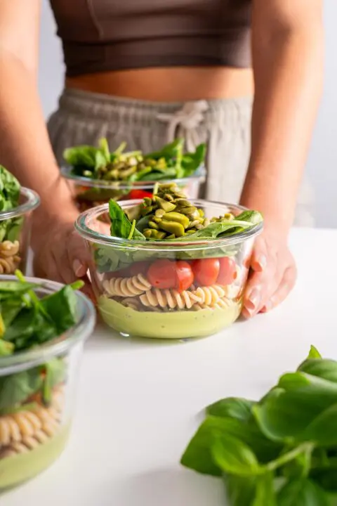 A person gently arranges four glass meal prep containers on a table, each filled with layers of creamy pistachio pesto, spelt pasta, cherry tomatoes, greens, and sprinkled with pumpkin seeds, ready for the week ahead.