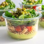 Progression of meal prep for Pistachio Pesto Pasta Salad, with the ingredients layered in glass containers; starting with a green pesto base, then adding beans, pasta, and tomatoes.