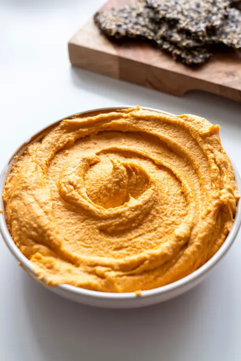Close-up of a bowl of red lentil hummus with an enticing swirl pattern on top with crackers on a chopping board in the background.