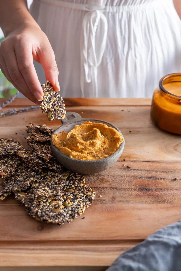 Hand holding a seeded cracker over a wooden table with hummus and a candle in the background.