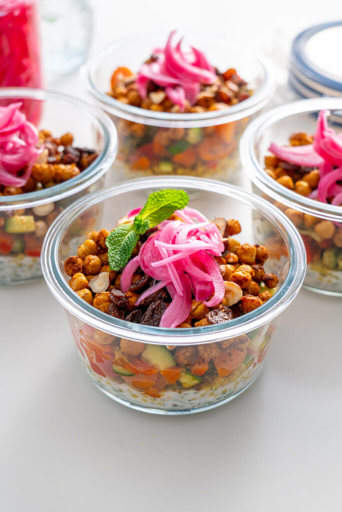 Các thùng chứa bữa ăn bằng thủy tinh chứa đầy salad couscous, đậu xanh nướng và trang trí bằng lá bạc hà và hành tây ngâm.