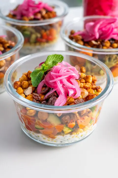 Close-up of a glass meal prep container with layers of dressing, roasted veggies and chickpeas with pickled onions on top.