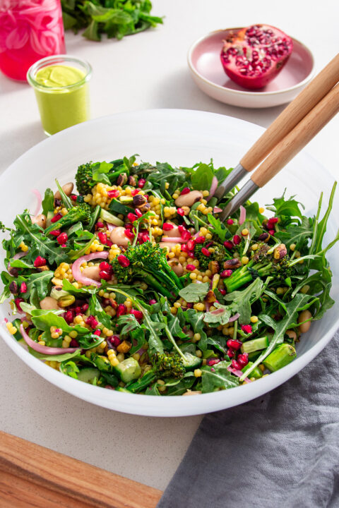 A large bowl of grilled broccolini and pomegranate salad with rocket and couscous on a white table, complete with serving utensils and fresh ingredients in the background.