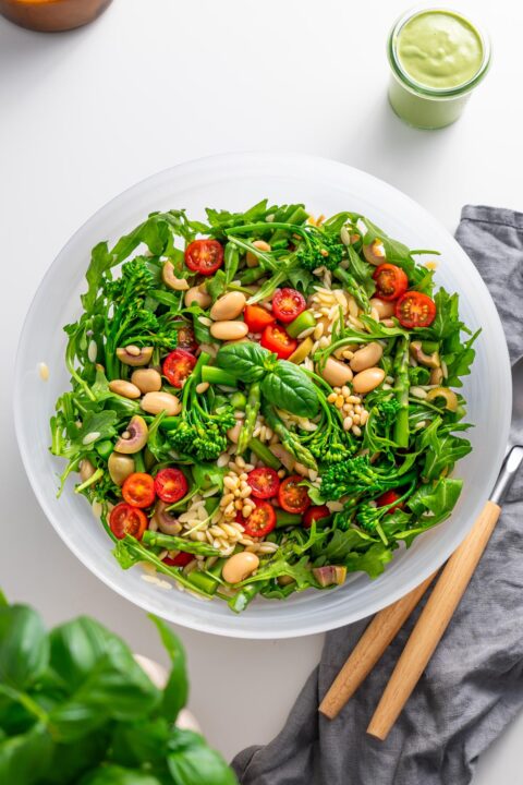 Top view of freshly prepared Pesto Orzo Salad in a white bowl on a kitchen counter, featuring arugula, broccolini, and cherry tomatoes.