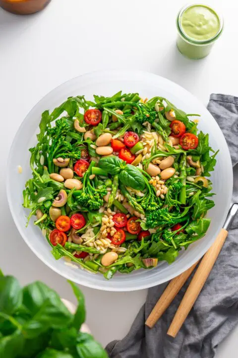 Top view of freshly prepared Pesto Orzo Salad in a white bowl on a kitchen counter, featuring arugula, broccolini, and cherry tomatoes.