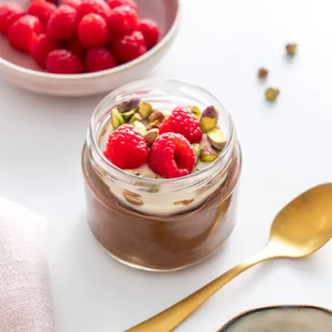 Cacao chia pudding jar topped with fresh raspberries and pistachios, with a spoon nearby.