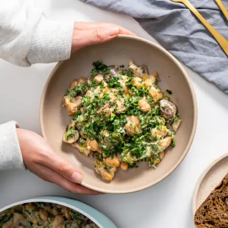 Sarah Cobacho’s hands holding creamy miso butter beans with kale next to toasted bread on a table.