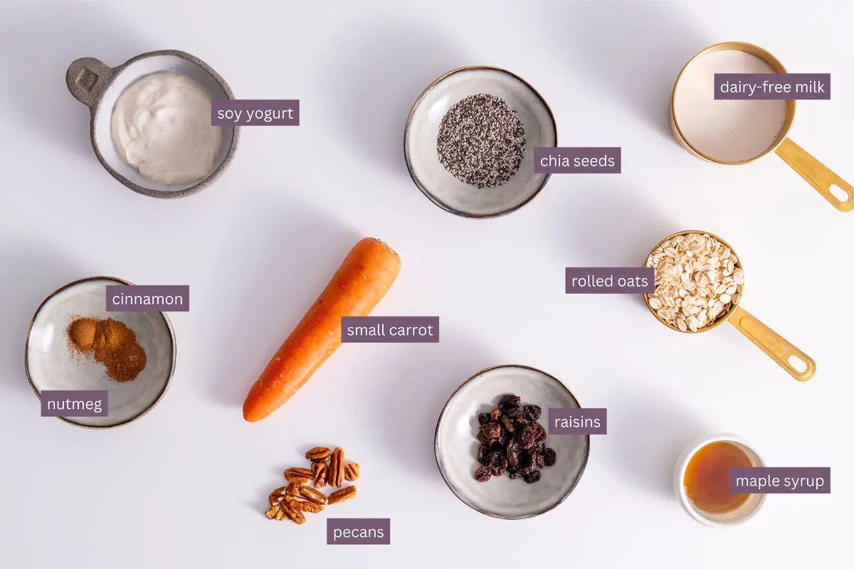 Vibrant ingredients for carrot cake overnight oats on a white surface, including chia seeds, oats, raisins, and a small carrot.