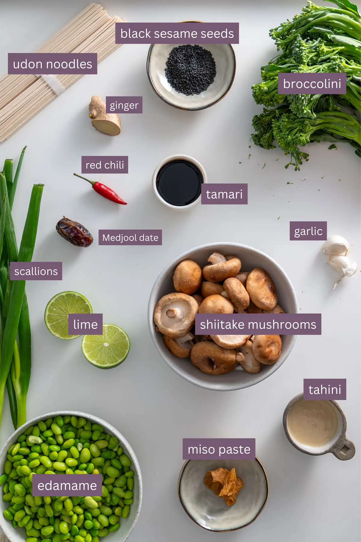 Vibrant ingredients for high-protein udon noodles, including edamame, broccolini, mushrooms, and seasonings, displayed on a white surface.