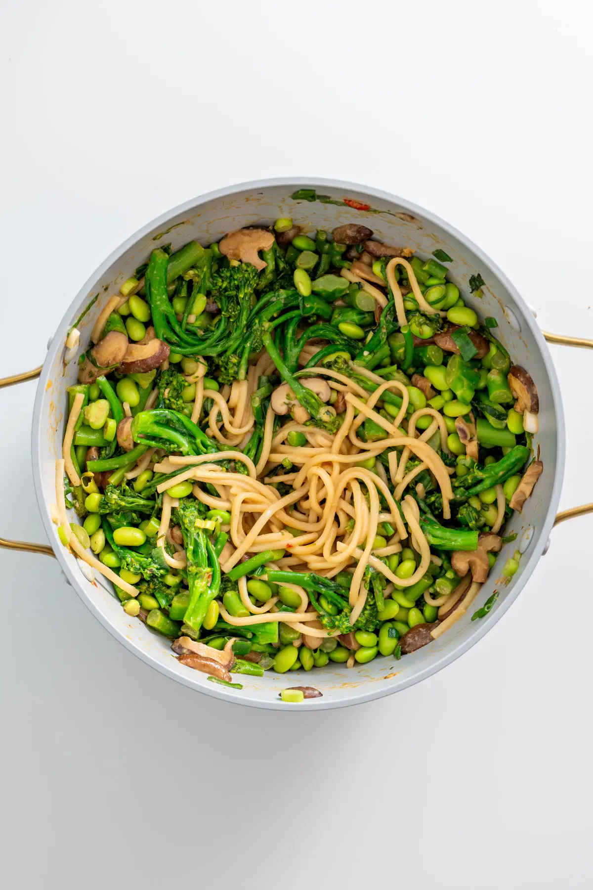 Close-up of a pot filled with high-protein udon noodles, broccolini, shiitake mushrooms, and edamame on a white surface.