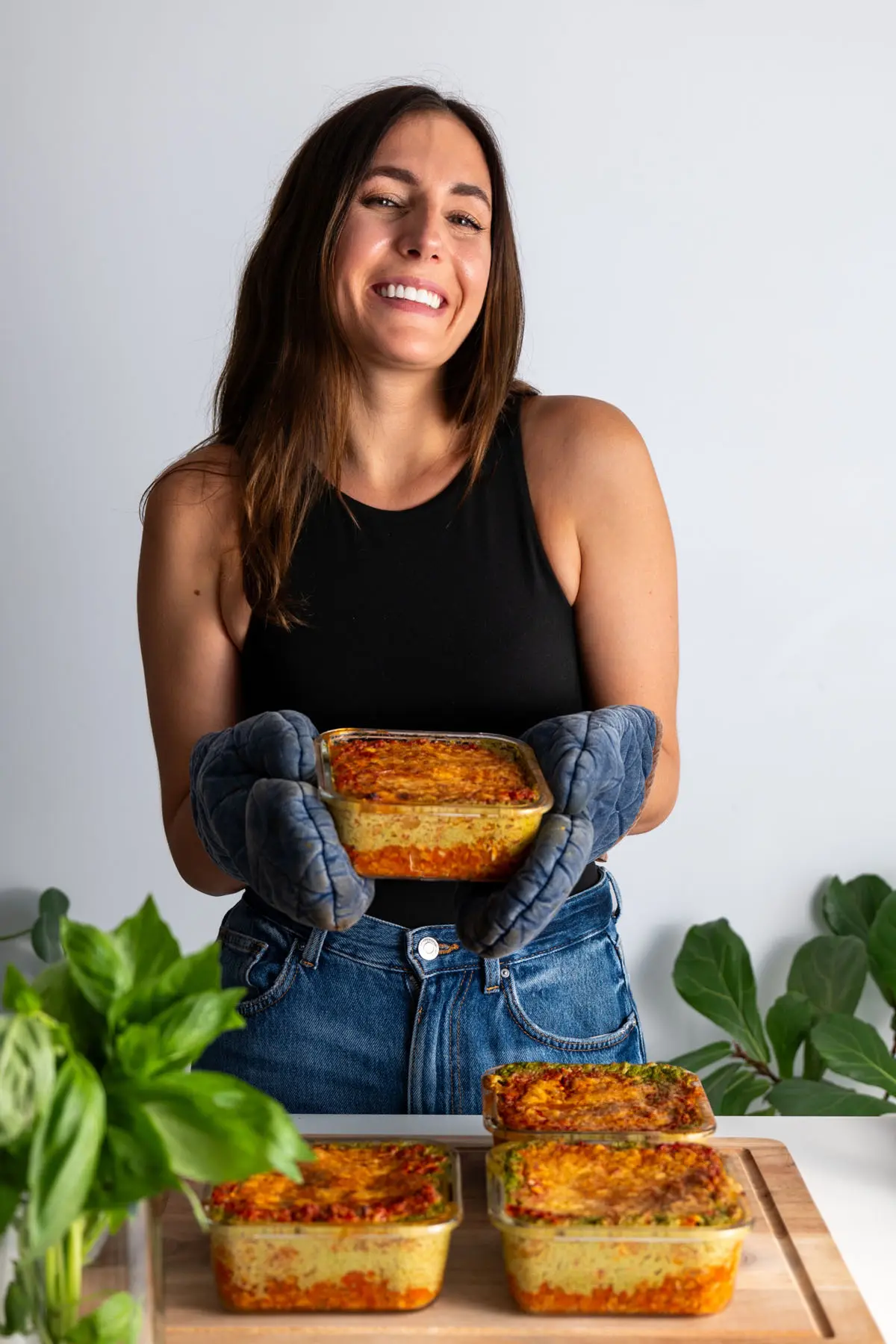 Sarah Cobacho smiling and holding a container of high-protein vegan lasagna, surrounded by meal prep containers on a wooden board.