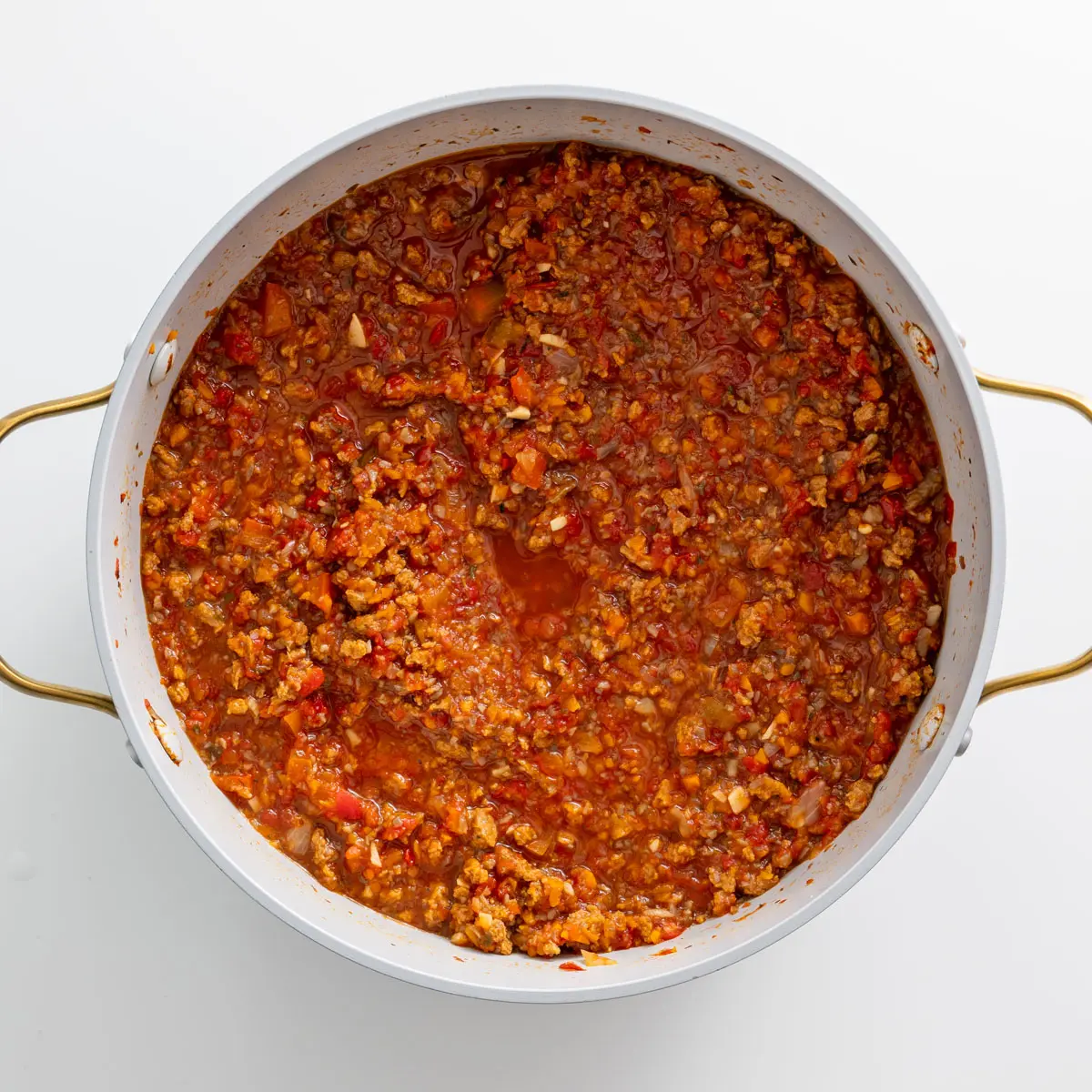 Rich and chunky vegan bolognese sauce in a white pot with golden handles on a white background.