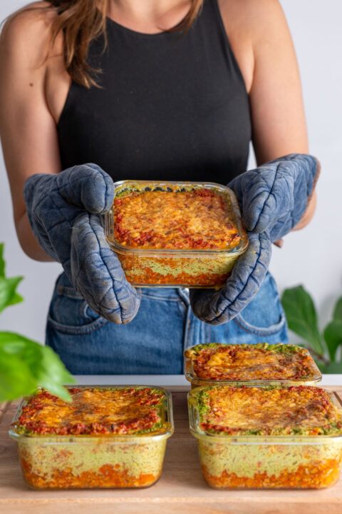 Sarah Cobacho holding a glass meal prep container filled with golden baked high-protein vegan lasagna.