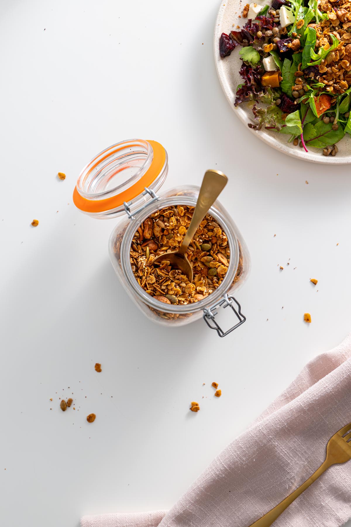 Lọ granola mặn tự làm với một cái muỗng vàng, được đặt trên một bề mặt trắng với một món salad trong nền.