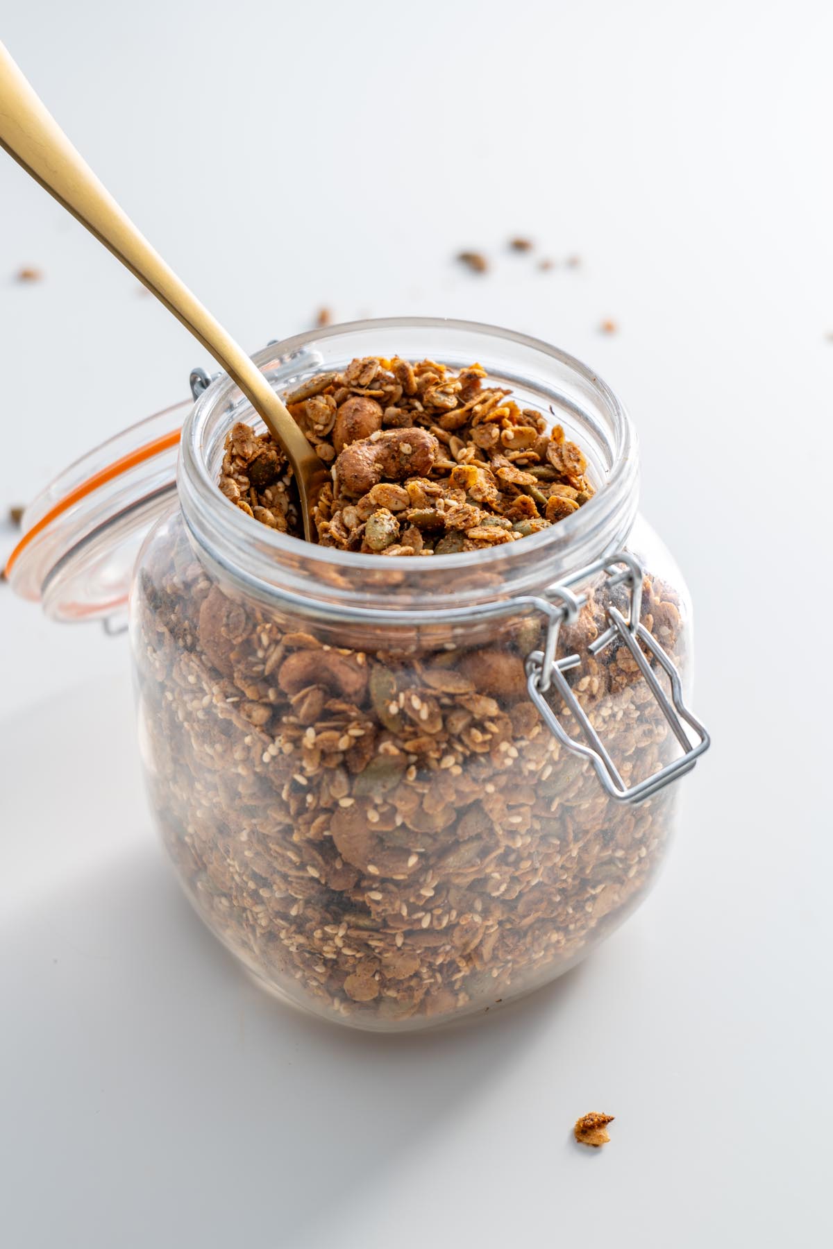 Một lọ granola mặn mới nướng với một cái muỗng vàng nằm bên trong, được đặt trên một bề mặt trắng sạch.