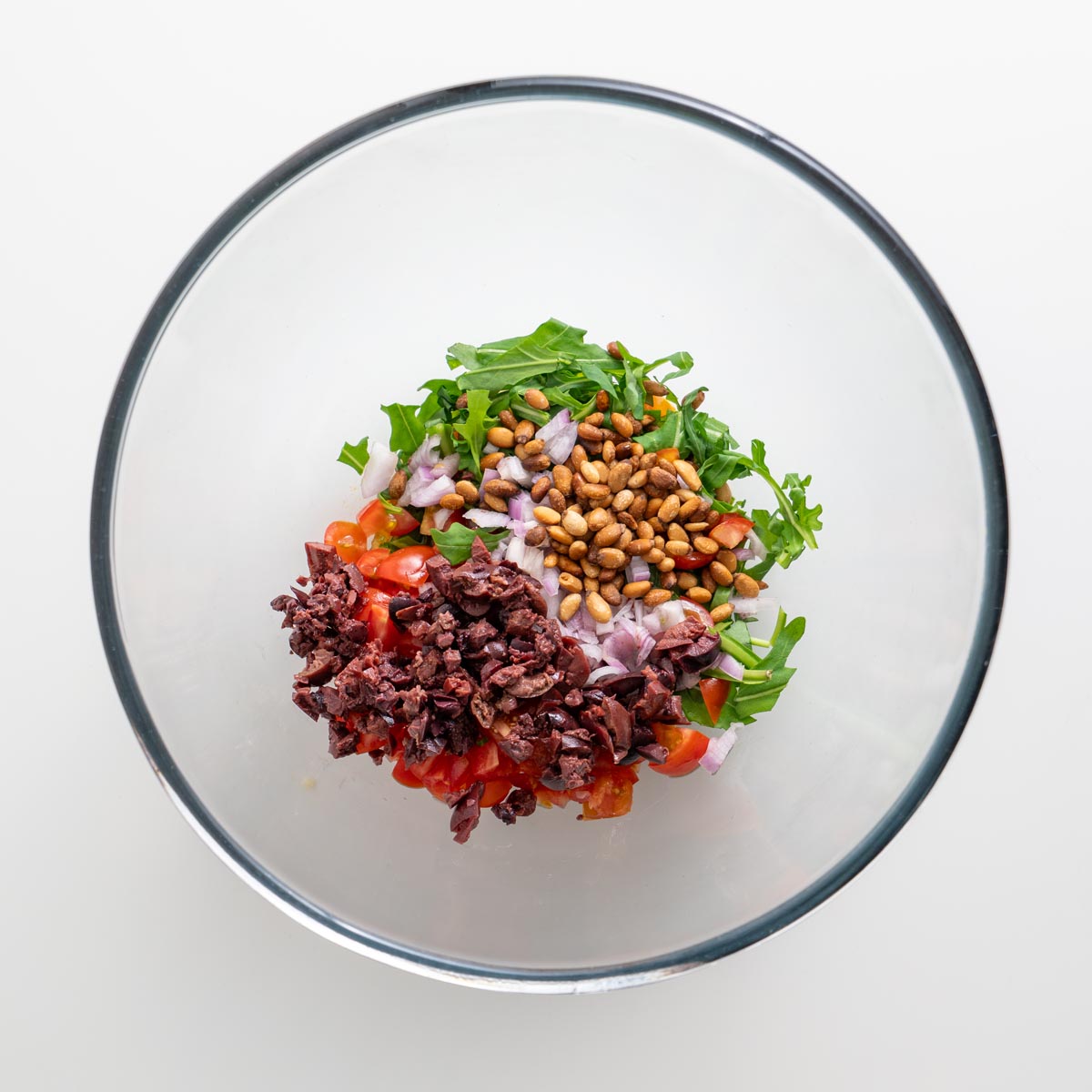 Olive mix ingredients in a bowl, including arugula, tomatoes, chopped olives, shallots, and toasted pine nuts
