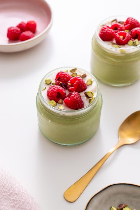 Jar of blended matcha chia pudding topped with dairy-free yogurt, fresh raspberries, and pistachios on a white surface.