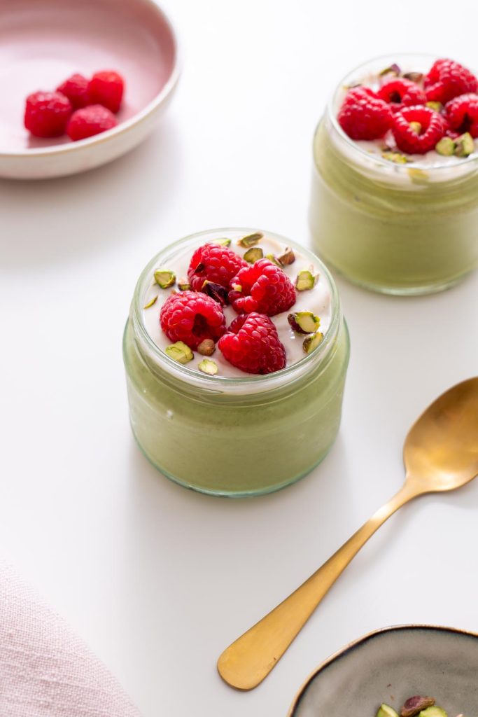 Jar of blended matcha chia pudding topped with dairy-free yogurt, fresh raspberries, and pistachios on a white surface.