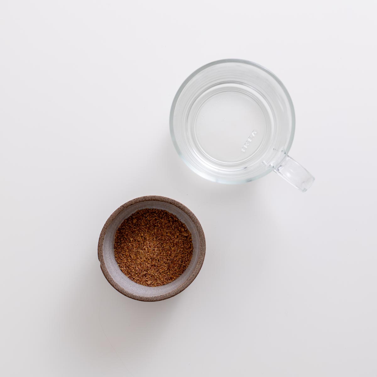 Cup of water next to a small bowl of ground flaxseed on a white background