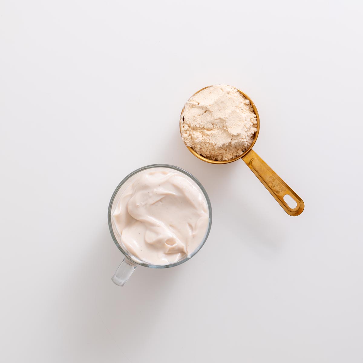 Glass cup of thick soy yogurt next to a gold measuring cup of vanilla protein powder on a white surface