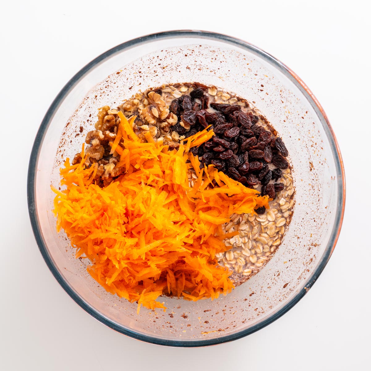 Glass mixing bowl with oats, shredded carrots, raisins, and walnuts before mixing