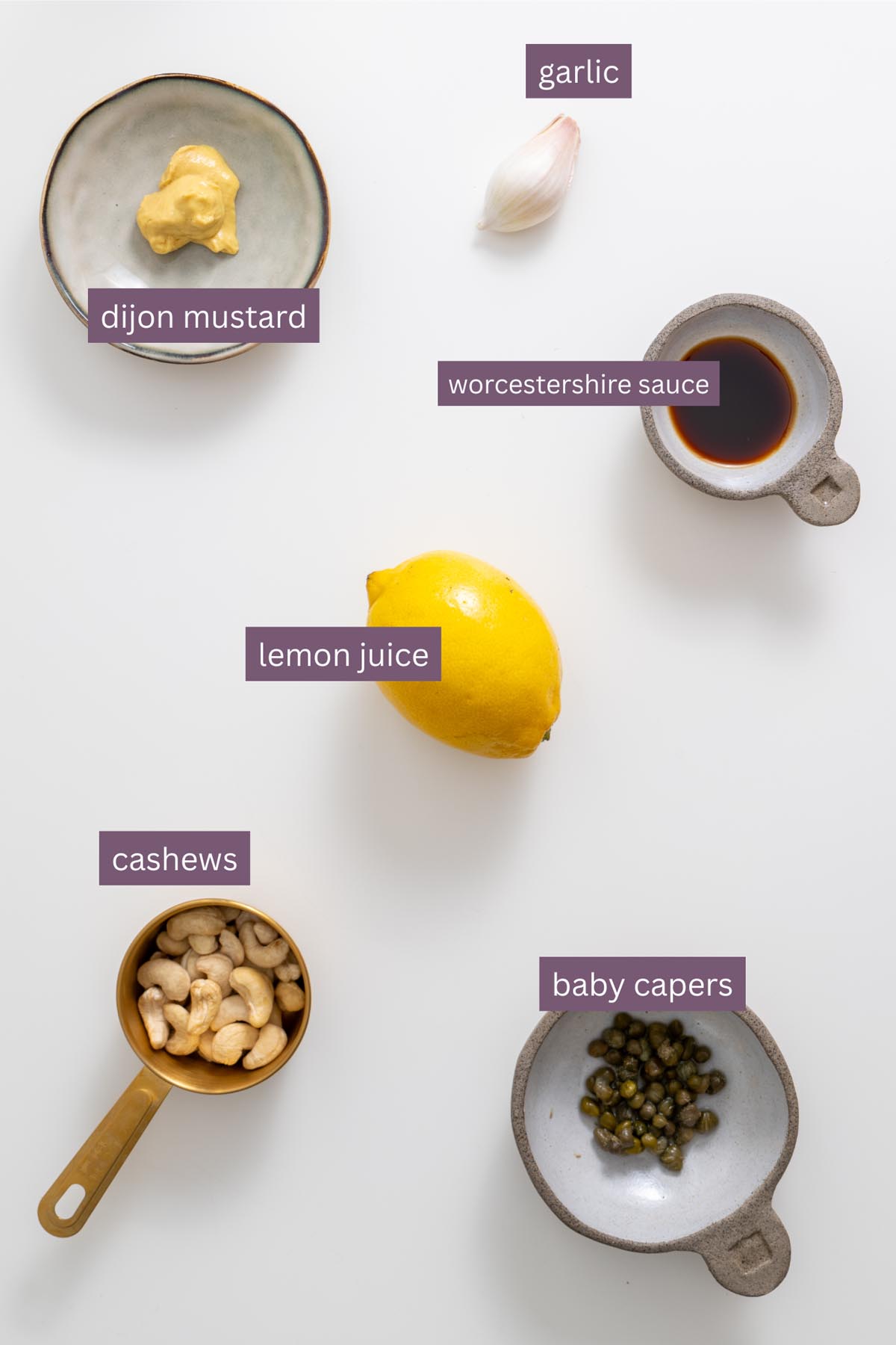 Vibrant ingredients for a vegan Caesar salad dressing on a white surface including garlic, lemon, cashews, capers, Dijon mustard, and Worcestershire sauce.