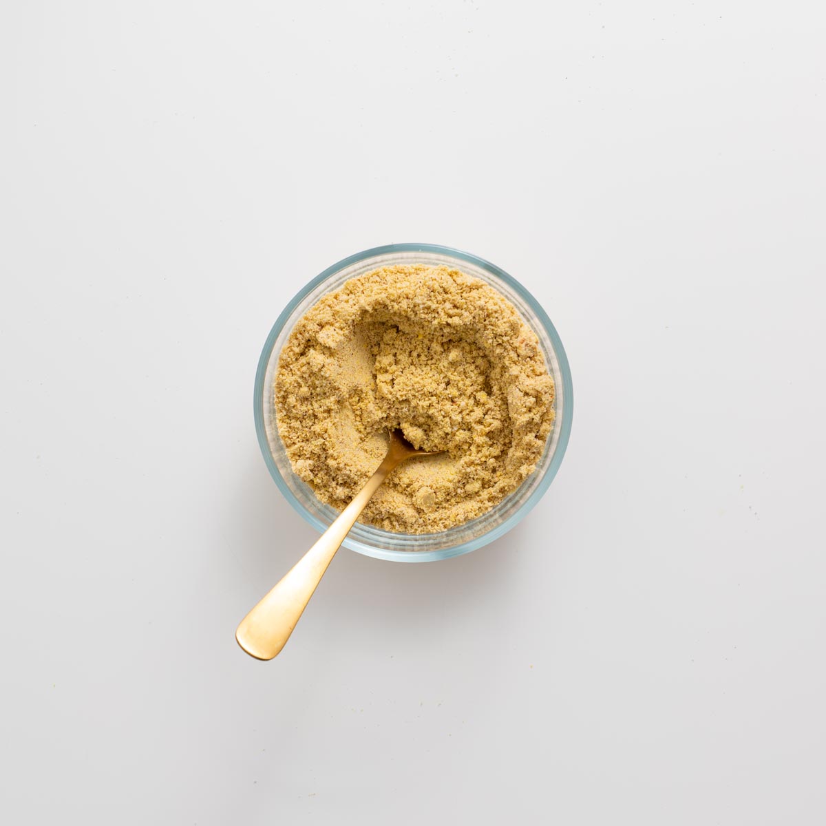 Homemade vegan parmesan in a glass bowl with a gold spoon on a white background.