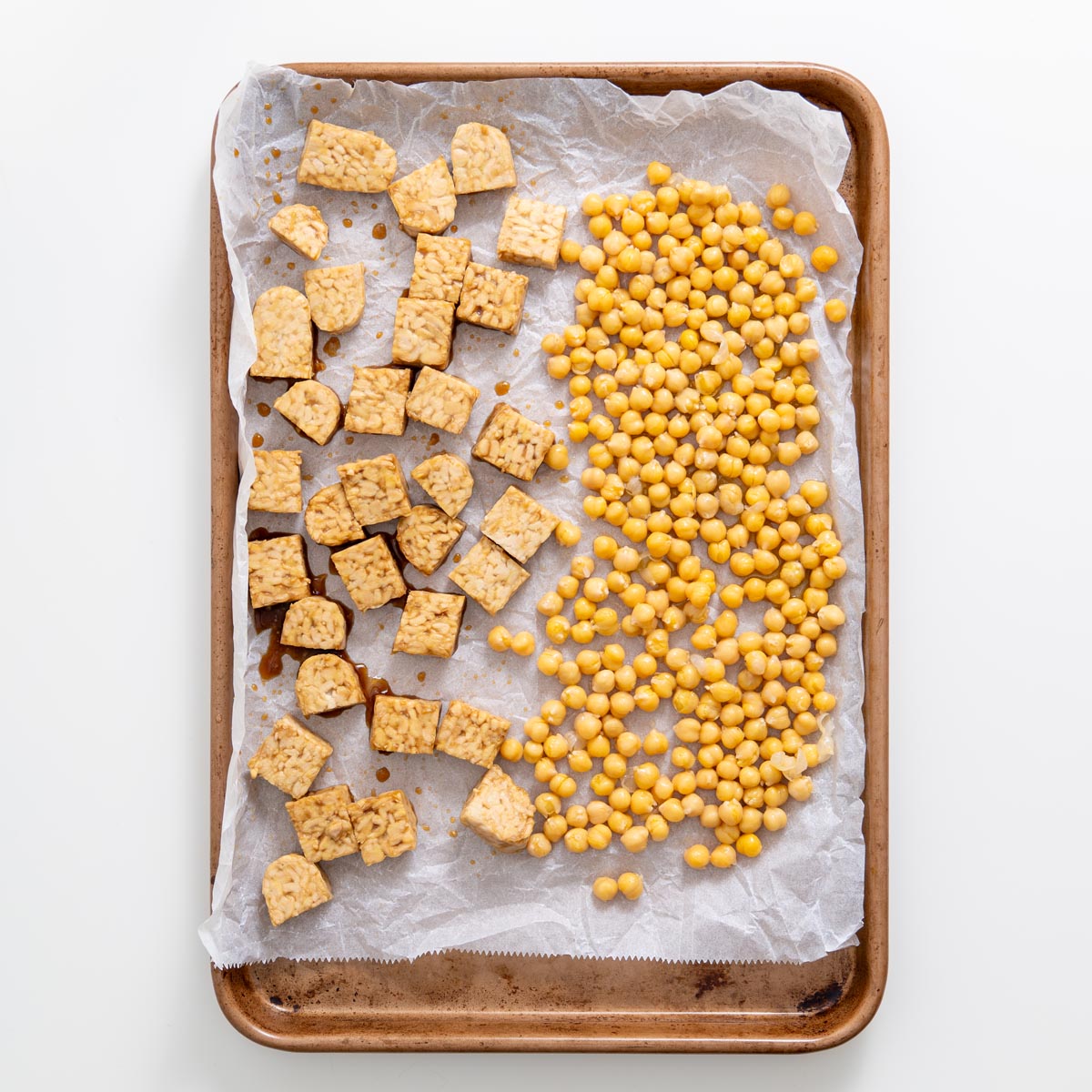 Baking tray with raw tempeh cubes and chickpeas before roasting on parchment paper.