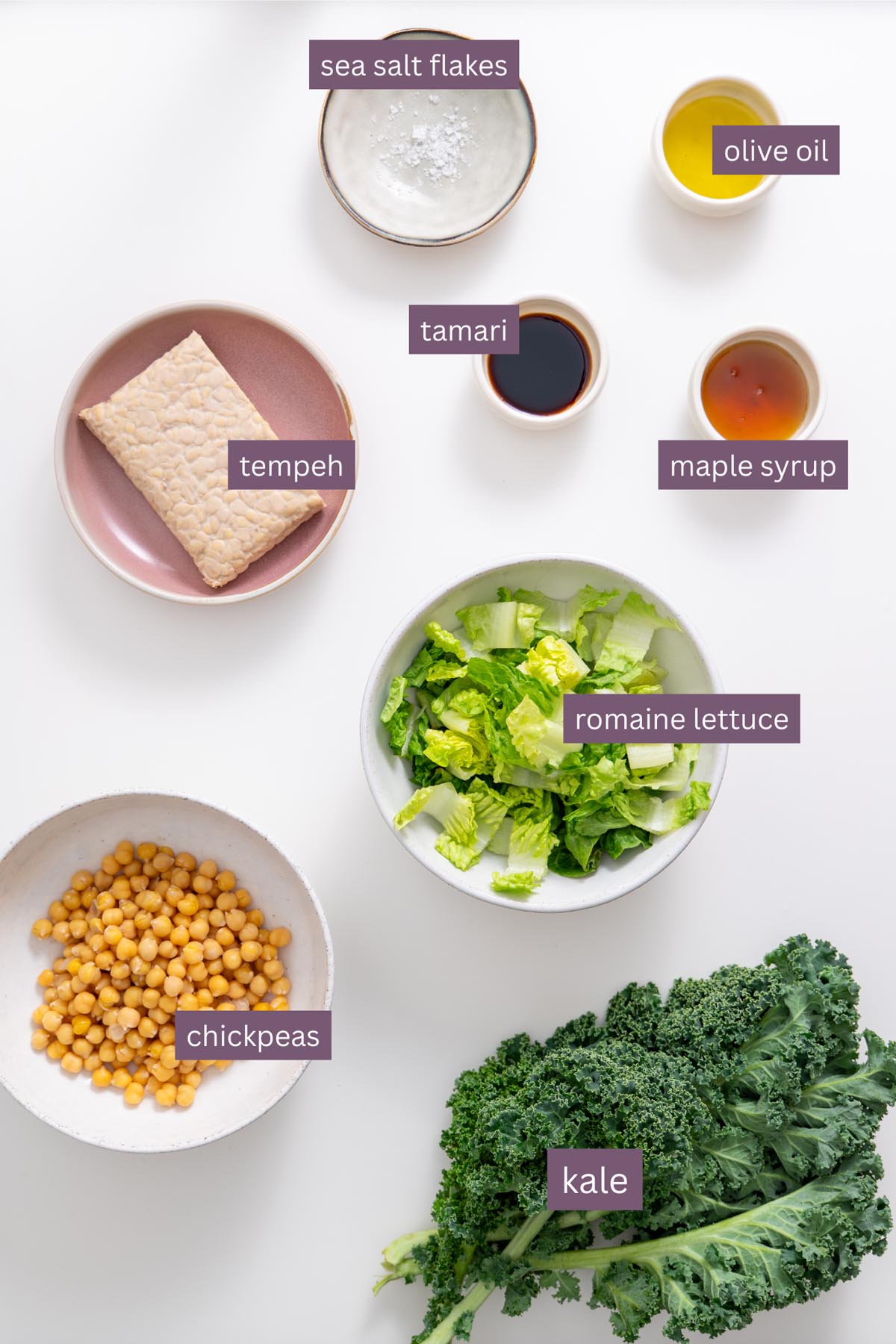 Fresh ingredients for a vegan Caesar salad on a white surface including romaine lettuce, kale, chickpeas, tempeh, tamari, maple syrup, olive oil, and sea salt flakes.