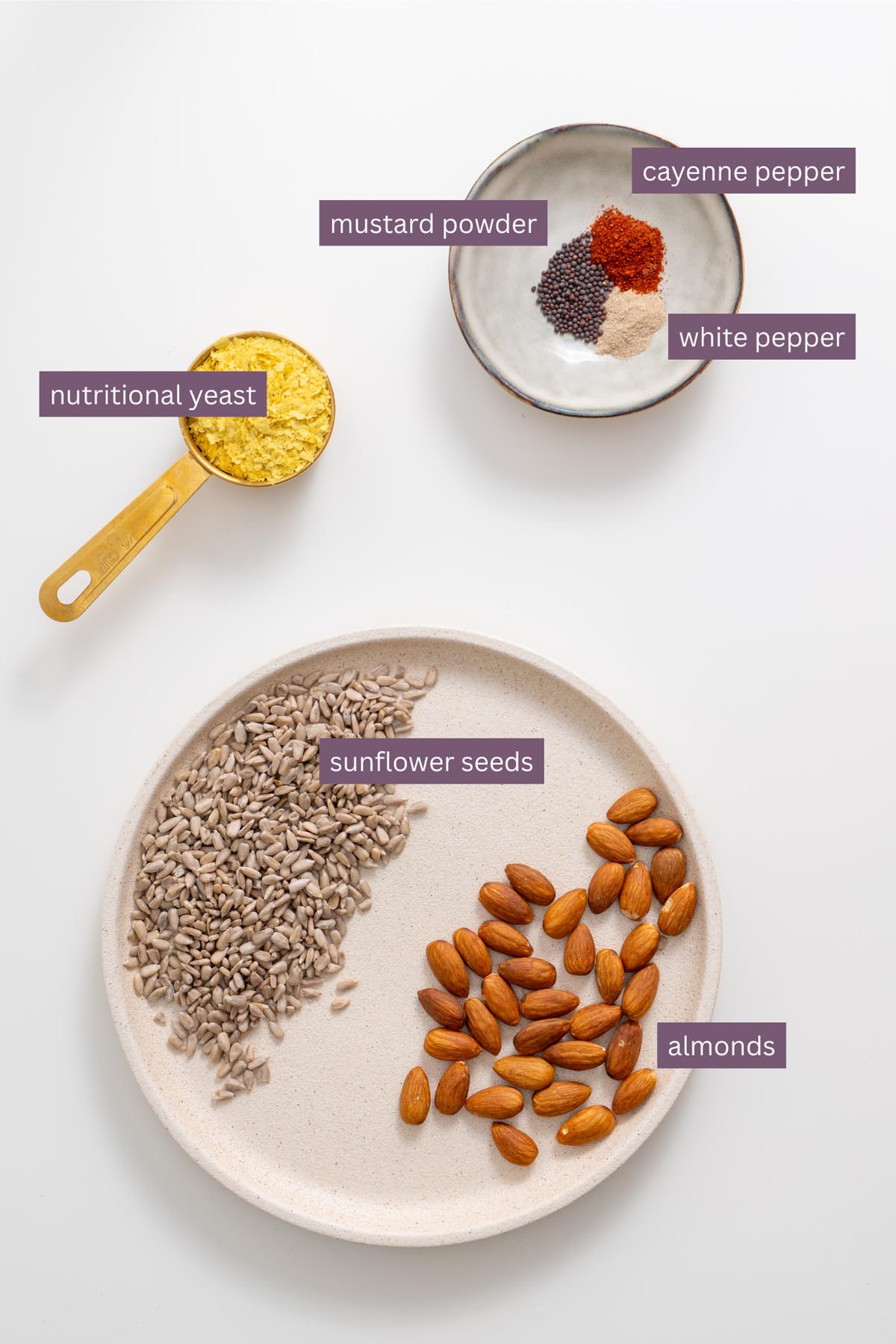 Vibrant ingredients for a vegan Caesar parmesan on a white surface including sunflower seeds, almonds, nutritional yeast, mustard powder, white pepper, and cayenne pepper.