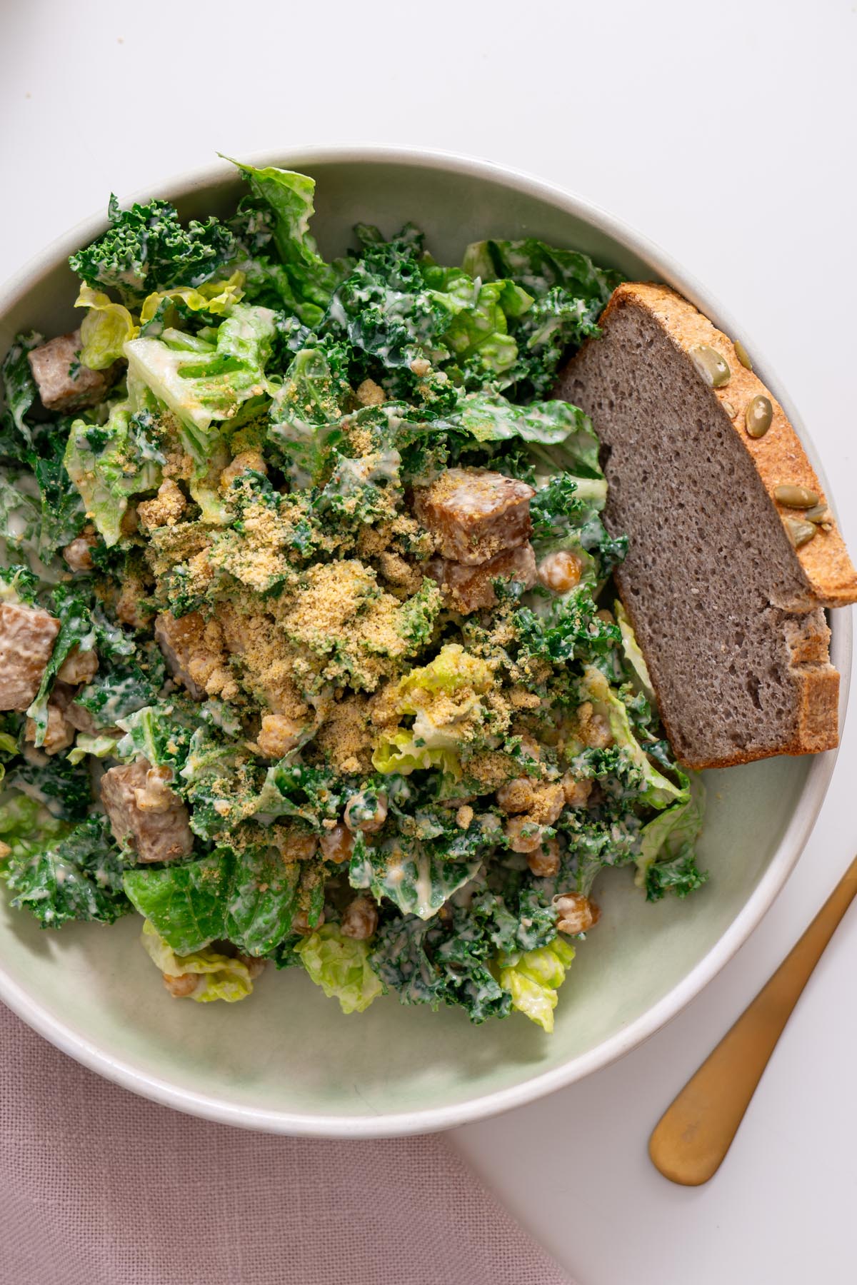 Bowl of vegan Caesar salad with romaine, kale, crispy tempeh, chickpeas, and parmesan topping served with bread.