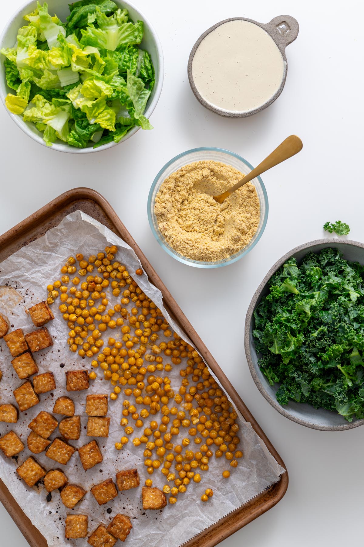 Prepared vegan Caesar salad ingredients including romaine, kale, baked tempeh, crispy chickpeas, dressing, and parmesan ready to be assembled.