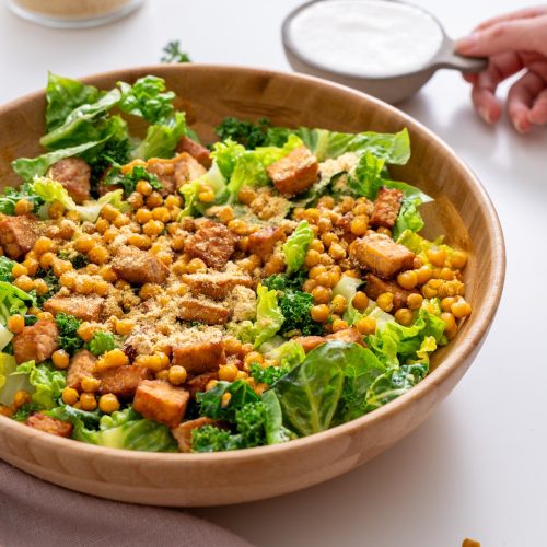 Sarah Cobacho holding a small bowl of creamy vegan Caesar dressing next to a salad with chickpeas, kale, romaine, and tempeh.