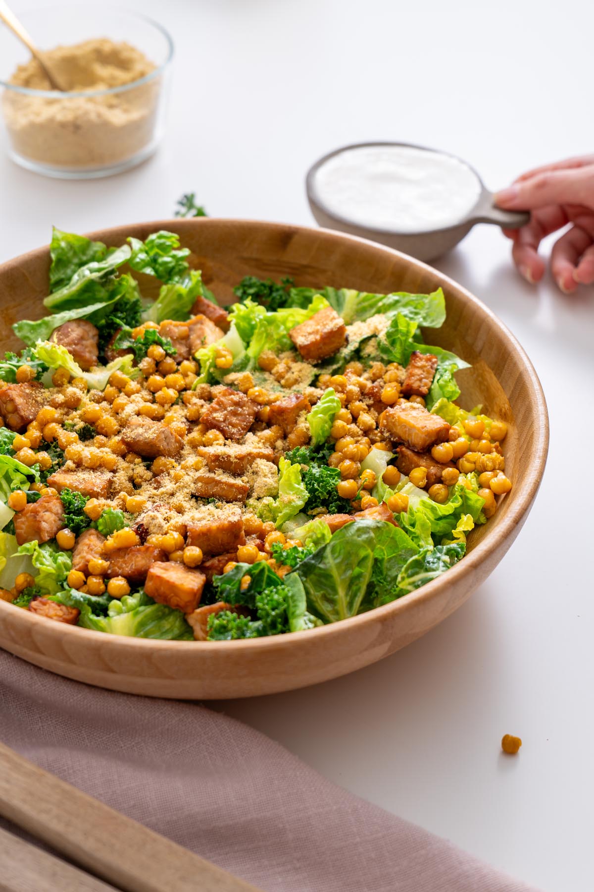 Sarah Cobacho holding a small bowl of creamy vegan Caesar dressing next to a salad with chickpeas, kale, romaine, and tempeh.